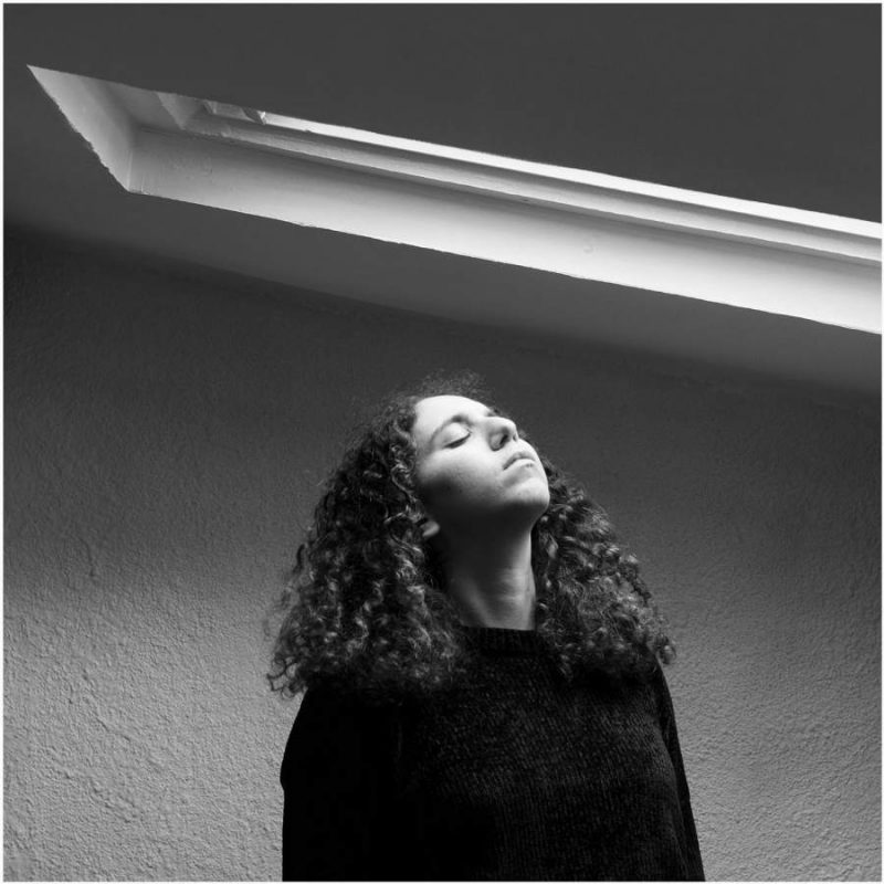 Portrait en noir et blanc de la photographe Maëva Benaiche en dessous d'une fenêtre Velux éclairée par la lumière du jour