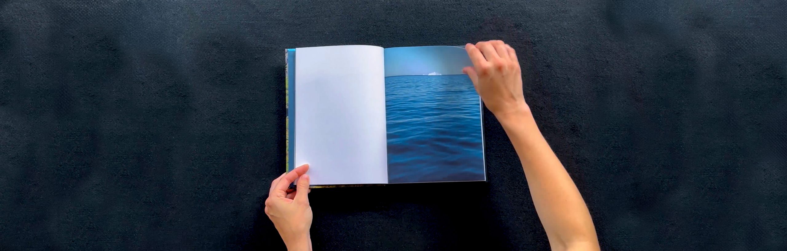 Visuel du livre de photographie et de poésie DANS LA GUEULE DU CIEL de Sandrine Cnudde publié par les Editions Light Motiv en 2018, en train d'être manipulé par une paire de mains blanches pour tourner les pages. L'image à l'arrêt représente une double page, la page de gauche restant blanche et celle de droite représentant la vue de la mer glacée avec un iceberg à l'horizon, à Tassilaaq au Groenland.